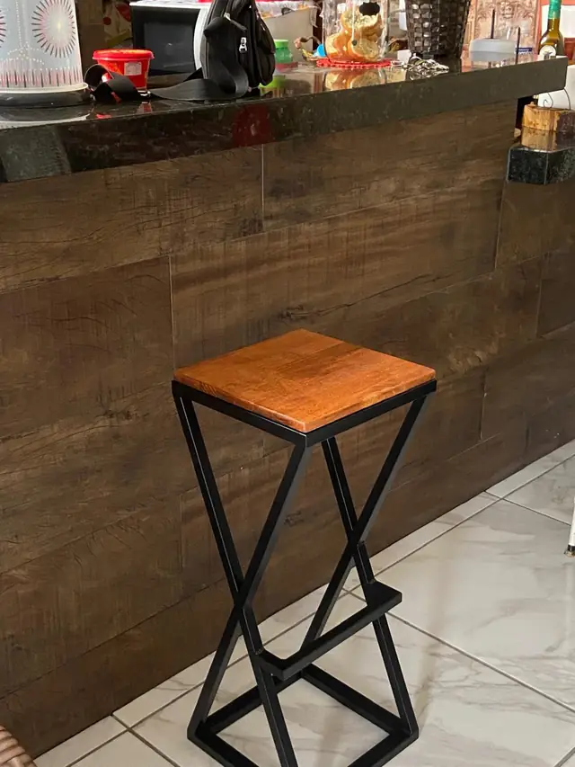 Banqueta alta de estrutura metálica preta em losango, com assento de madeira, encostada num balcão