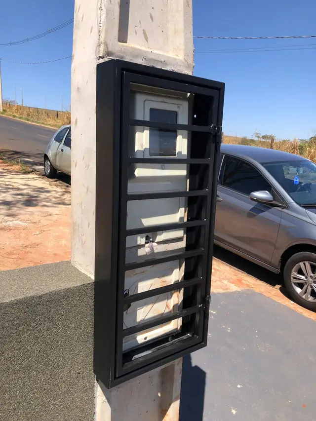 Caixa de proteção metálica preta instalada sobre o medidor de energia, no muro da calçada