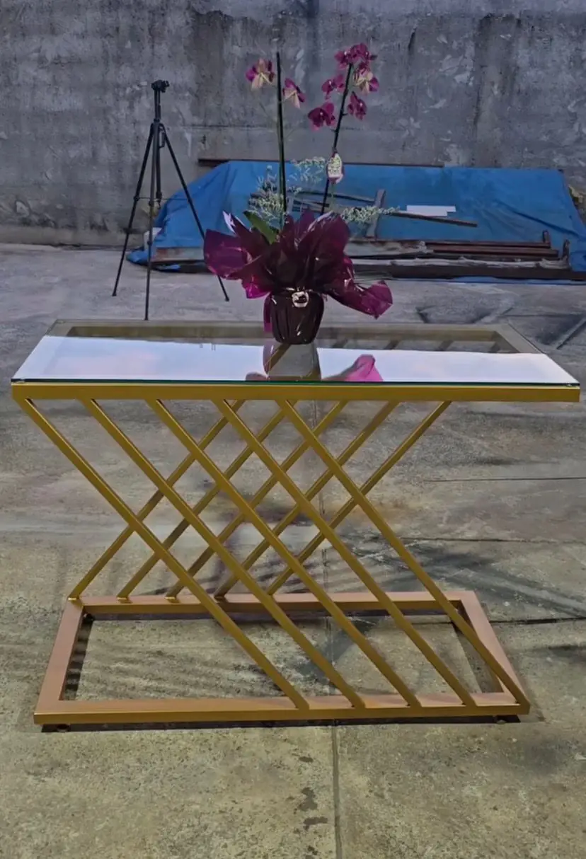 Mesa com base metálica dourada em grade de losangos e tampo de vidro, com um vaso de orquídea em cima